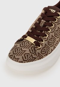 Brown patterned sneaker with textured fabric, golden eyelets, and a white rubber sole. Features a gold logo plate on the tongue.
