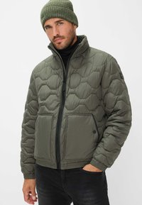 Polstret jakke i olivengrøn med quiltet design, frontlynlås og to store lommer. Har en stående krave og elastiske manchetter.