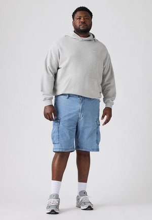 Un hombre luce una sudadera gris con capucha, unos pantalones cortos cargo vaqueros azules, calcetines blancos y zapatillas grises sobre un fondo de estudio sencillo.