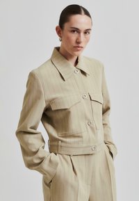 Beige randig jacka med bred krage, två framfickor och knappstängning. Tillverkad av ett mjukt tyg med skräddarsydd passform.