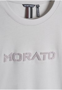Camiseta gris claro con cuello redondo, que presenta bordado texturizado en un gris ligeramente más oscuro que dice "MORATO". Etiqueta de ajuste regular incluida.