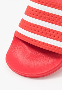 Röd slidesandal med texturerad ovandel och tre vita ränder, samt en slät, vadderad fotbädd. Tillverkad av slitstarkt syntetmaterial.