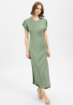 Vrouw draagt een vloerlang, saliegroen satijnen jurk met korte, gespleten mouwen en gouden sandalen met hoge hakken op een witte achtergrond.