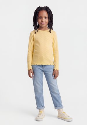 Młoda dziewczyna z zaplecionymi włosami stoi, ubrana w żółtą bluzkę z długim rękawem, jasnoniebieskie dżinsy, beżowe skarpetki oraz kremowe trampki.