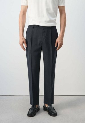 Uomo con pantaloni neri su misura, mocassini in pelle neri e camicia bianca a maniche corte, in piedi davanti a uno sfondo uniforme.