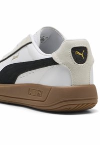 Sneaker en cuir blanc avec une bande en daim noir, un accent arrière en daim beige, un logo doré et une semelle en caoutchouc gomme. Design classique avec des textures contrastantes.