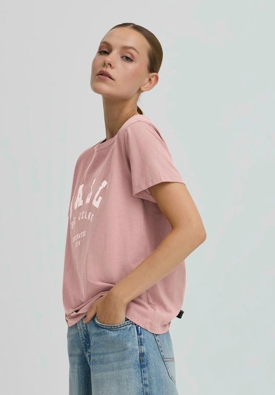 Žena s vlasmi stiahnutými dozadu, oblečená v ružovom tričku s bielym potlačou a svetlomodrých džínsoch, stojí s jednou rukou vo vrecku na svetlom pozadí.