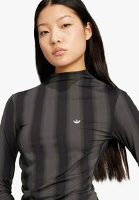 Giovane donna con lunghi capelli scuri indossa una maglia a collo alto a righe nere e grigio scuro con un piccolo logo argentato sul petto.