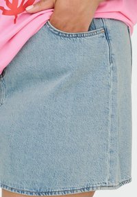 Mano en el bolsillo de una falda vaquera azul claro combinada con una camisa rosa, mostrando la textura del tejido y los detalles del bolsillo.