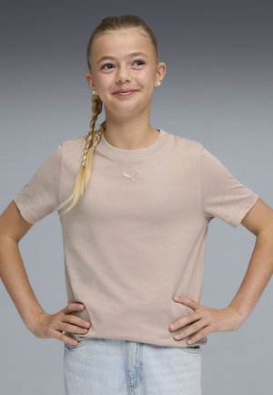 Niña joven con el cabello trenzado sonríe, con las manos en las caderas, vistiendo una camiseta beige de Puma y jeans azul claro sobre un fondo gris.