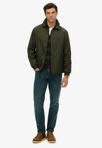 Zielona, wodoodporna kurtka zapinana na napy, z elastycznymi mankietami i wysokim kołnierzem. Nosi na sobie koszulę w kratę, w zestawieniu z niebieskimi dżinsami i brązowymi botkami.