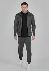 Grijze sportieve jas met rits over een zwart shirt, gecombineerd met grijze joggers en witte sneakers. Glad materiaal met een getailleerd ontwerp.