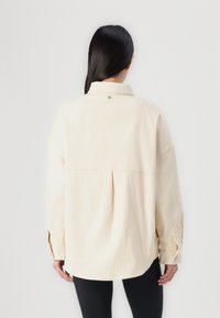 Chemise en velours côtelé beige avec un col, des manches longues et des poignets boutonnés. Comprend un empiècement au dos et un ourlet arrondi. Rayures verticales texturées.