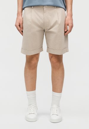 Homme portant un short beige retroussé, des chaussettes blanches montantes et des baskets blanches, debout devant un fond clair uni.