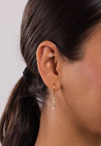 Goldene Creolenohrringe mit rechteckigen und runden klaren Edelsteinen, die an einem kleinen goldenen Verschluss hängen und eine Pferdeschwanzfrisur ergänzen.