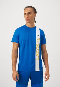 Modra majica s kratkimi rokavi z belim navpičnim pasom, na katerem je napis "LACOSTE" v rumeni barvi, izdelana iz gladkega materiala. Lacoste logotip na prsih.