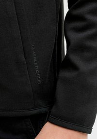 Juoda sportinė palaidinė su tekstūruota medžiaga, šonuose su logotipo akcentu ir siūlelio apačia, pabrėžianti aptemptą dizainą ir rankovių manžetus.