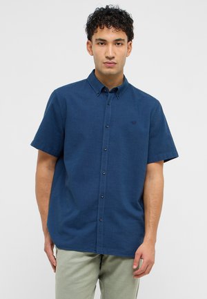 Mężczyzna z kręconymi włosami, ubrany w granatową, zapinaną na guziki koszulę z krótkim rękawem oraz jasnozielone spodnie, stojący na jednolitym tle.