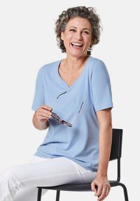 Lichtblauw kortmouwen top met een V-hals, voorzien van kleine decoratieve knopen. De drager houdt een zonnebril vast, zittend op een zwarte stoel.