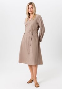 Taupe jurk met een V-hals en een strik taille. Lange mouwen met oogjesdetails. Gladde stof met een knie-lange snit en een ontspannen pasvorm.