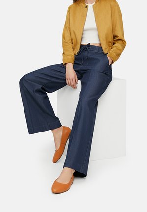 Weite marineblaue Hosen mit einer strukturierten Oberfläche, kombiniert mit einer senfgelben Wildlederjacke und orangefarbenen Ballerinas. Einfaches Design mit minimalen Akzenten.