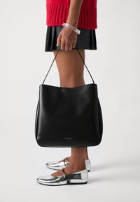 Black leather tote bag with a smooth texture, featuring a rounded top and a gold logo at the base. Worn with a red sweater and silver flats.
