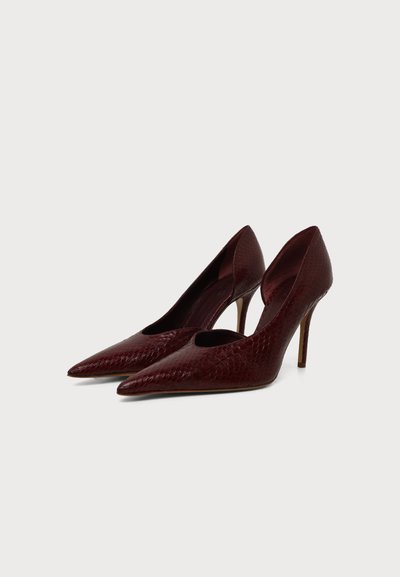 Paire d'escarpins en cuir texturé rouge foncé avec bouts pointus et talons aiguilles fins sur un fond clair uni.