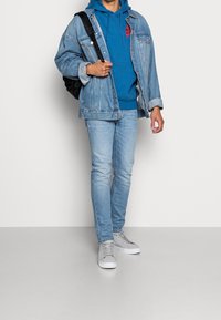 Giubbotto di jeans, felpa blu con logo rosso, jeans azzurri e sneakers grigie. Zaino nero indossato a spalla.