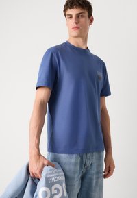 Zils īsām piedurknēm T-krekls, izgatavots no gludas kokvilnas, ar mazu logotipu kreisajā krūtī un valkāts ar gaišiem džinsu biksēm.