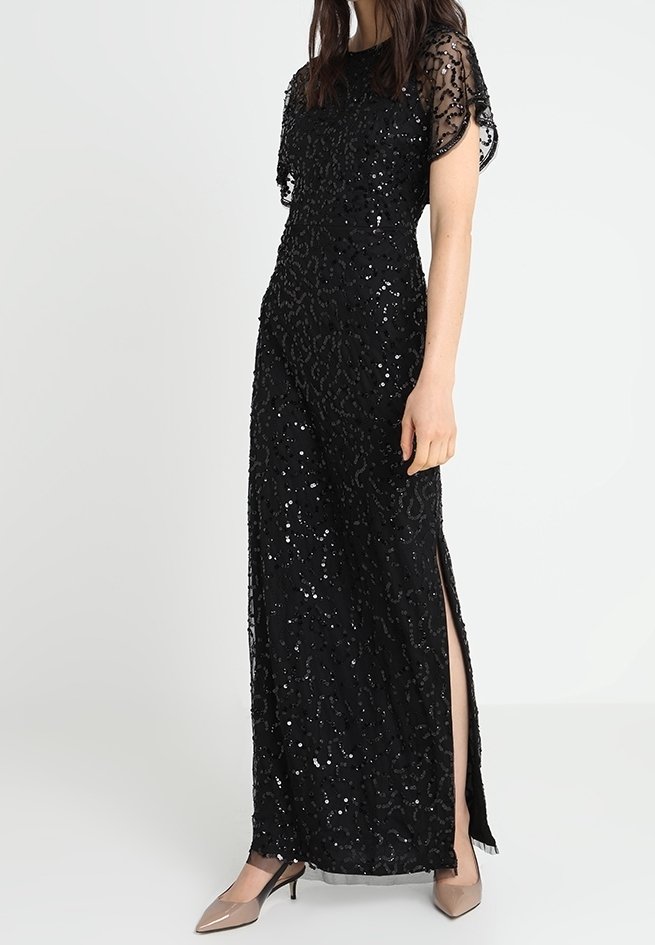 Robe noire longue jusqu'au sol avec des manches courtes, ornements de sequins, tissu texturé et fentes sur les côtés. Portée avec des talons beiges.