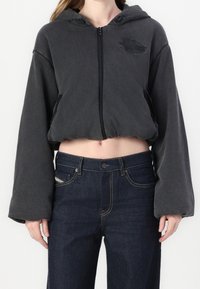 Sudadera corta con cremallera en tela gris oscuro, con capucha, mangas anchas y un sutil logo en el pecho. Combinada con vaqueros oscuros de mezclilla.