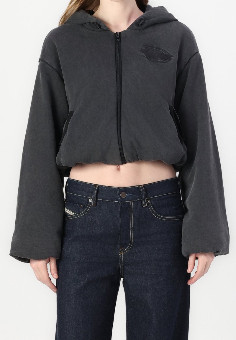 Sudadera corta con cremallera en tela gris oscuro, con capucha, mangas anchas y un sutil logo en el pecho. Combinada con vaqueros oscuros de mezclilla.