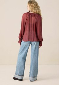 Blouse bordeaux transparente avec col à volants et manches longues, associée à un jean clair à jambes larges avec un ourlet retroussé.