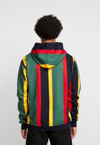 Persona con pelo rizado, vistiendo una sudadera verde con rayas verticales rojas, amarillas y negras, de pie mirando hacia atrás frente a un fondo liso.