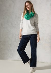 Grauer Strickpulli, lockere Passform. Grüner, leichter Schal, um den Hals drapiert. Dunkelblaue, weit geschnittene Jeans mit Kontrastnähten. Weiße Sneaker.