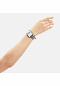 Silverfärgad metallklocka med fyrkantig urtavla, vit skiva och metallnätband, som kompletteras med en tunn kedjad detalj på handleden.