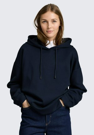 Junge Frau mit hellbraunen Haaren, die einen schwarzen Kapuzenpullover und blaue Jeans trägt, die Hände in den Taschen, schaut mit neutralem Gesichtsausdruck nach vorne.