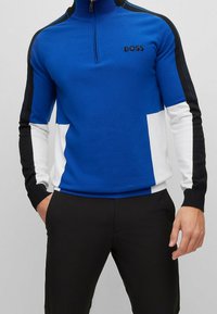 Hombre con una camiseta de manga larga con cremallera en azul, blanco y negro con el logo "BOSS" en el pecho y pantalones negros, sobre un fondo gris.