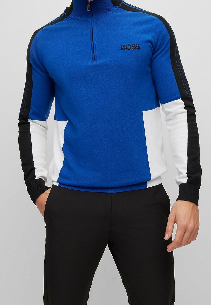 Hombre con una camiseta de manga larga con cremallera en azul, blanco y negro con el logo "BOSS" en el pecho y pantalones negros, sobre un fondo gris.