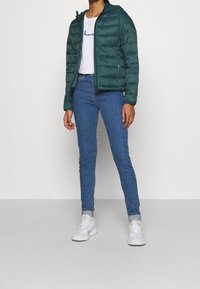 Grön puffjacka med dragkedja och huva, tillsammans med blåa tajta jeans och vita sneakers. Slät tygtextur, enkel design.