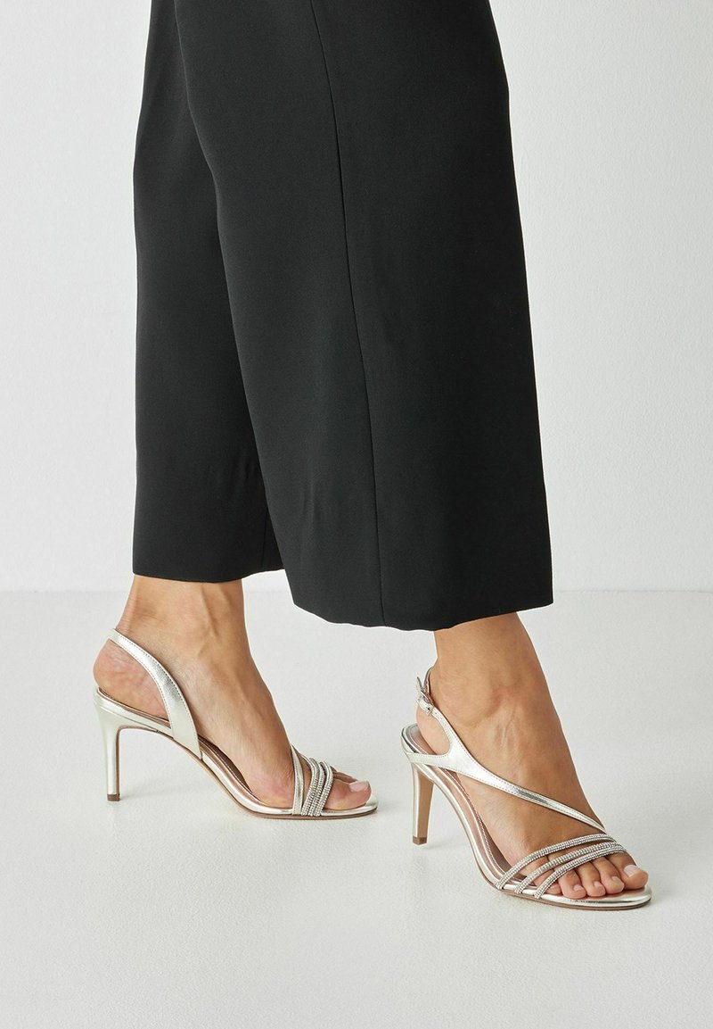 Pieds portant des sandales à talons hauts argentées avec des brides en strass, associées à un pantalon large noir raccourci sur un sol blanc.