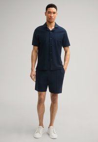 Marineblauw kortemouwenshirt met een subtiel gestructureerd patroon, aangevuld met bijpassende shorts, gedragen met witte sneakers. Knopen zijn zichtbaar.