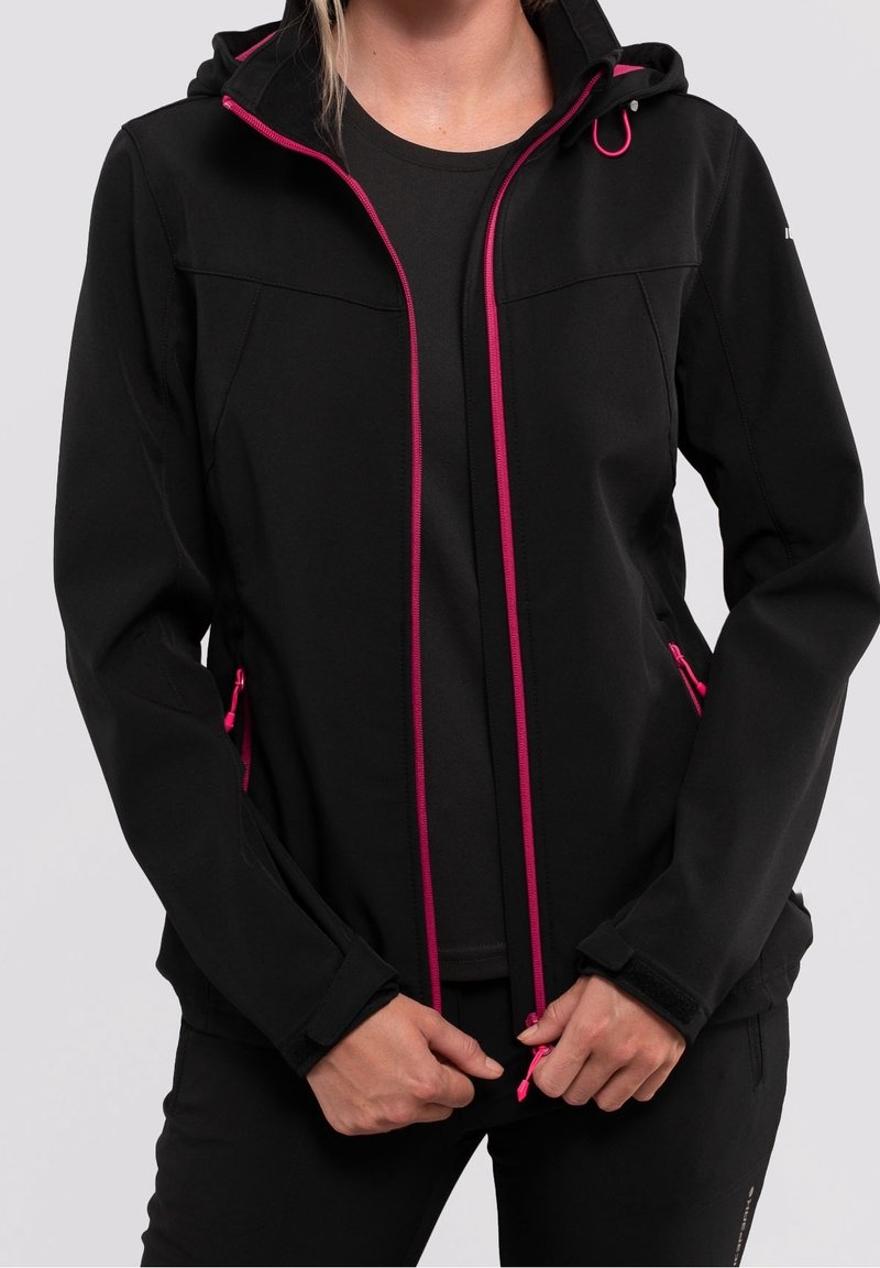 Femme portant une veste noire à capuche avec des fermetures éclair et des détails roses, tenant le curseur de la fermeture éclair à l'avant.