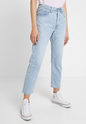 Femme portant un jean droit taille haute bleu clair et des baskets blanches montantes, debout devant un fond uni.