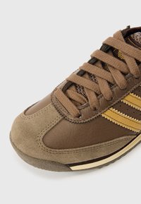 Καφέ αθλητικό παπούτσι από δέρμα και σουέντ, με μπεζ λεπτομέρειες, υφασμένες κορδόνια και τρεις κίτ�ρινες ρίγες στην πλευρά.