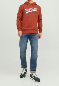 Felpa color ruggine con testo bianco, jeans blu e sneakers verdi. La felpa presenta una tasca frontale e cordini regolabili.