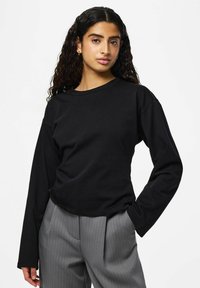 Chemise noire à manches longues avec un col rond et une coupe décontractée, en tissu lisse. Associée à un pantalon gris à fines rayures.
