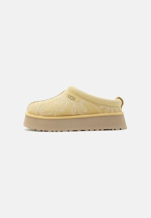 Chaussure UGG beige à enfiler avec semelle épaisse, couture florale et logo sur le côté, conçue pour une utilisation décontractée.