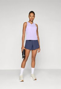 Helelilla varrukateta pluus ümarate kaelustega, kombineeritud tumesiniste pükstega. Modell hoiab musta joogipudelit, kannab valgeid sokke ja beeže tossusid.