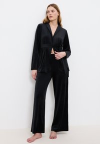 Zwart velours pyjama set met een wikkeltop met strikceintuur en een broek met wijde pijpen. Zachte textuur met twee zakken aan de voorkant van de top.
